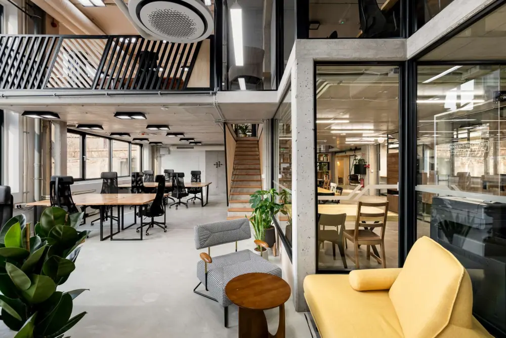 Open coworking area at Puzl CowOrKing with shared desks, glass meeting rooms, plants and a yellow sofa